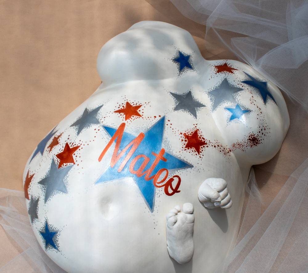 Escultura de barriga en yeso pintada con estrellas azules y rojas y el nombre Mateo, con el pie y la mano del bebé.